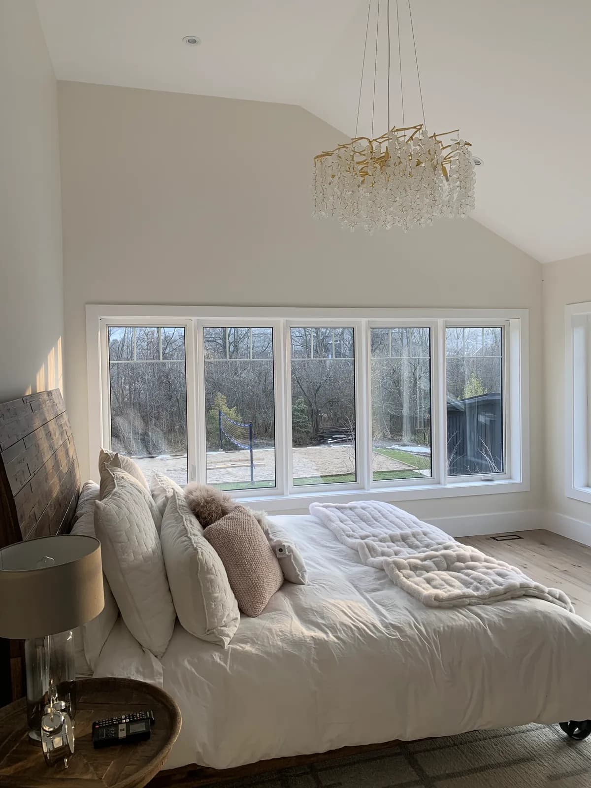 A bedroom in a Blue Mountain chalet with a vaulted painted ceiling, beaded chandelier and a wall of windows above layered cream bedding.