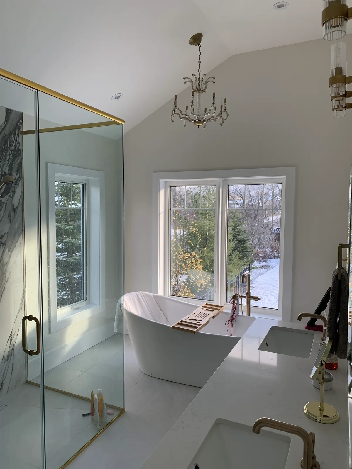 A Blue Mountain chalet ensuite with a freestanding white tub beside a window, a glass shower with brass framing, and a chandelier overhead.