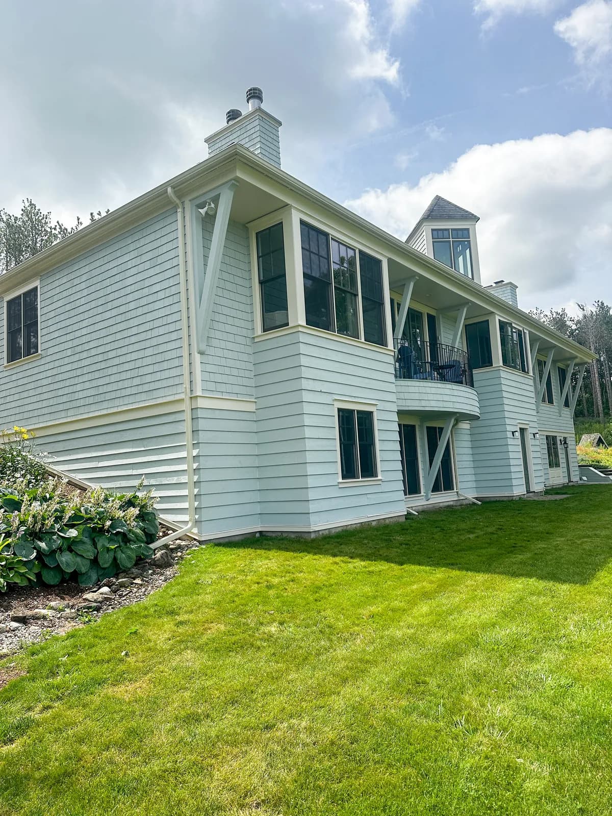 The side elevation of a Creemore home, its cedar shakes stained pale sage with crisp white trim and black window frames above a mown lawn.