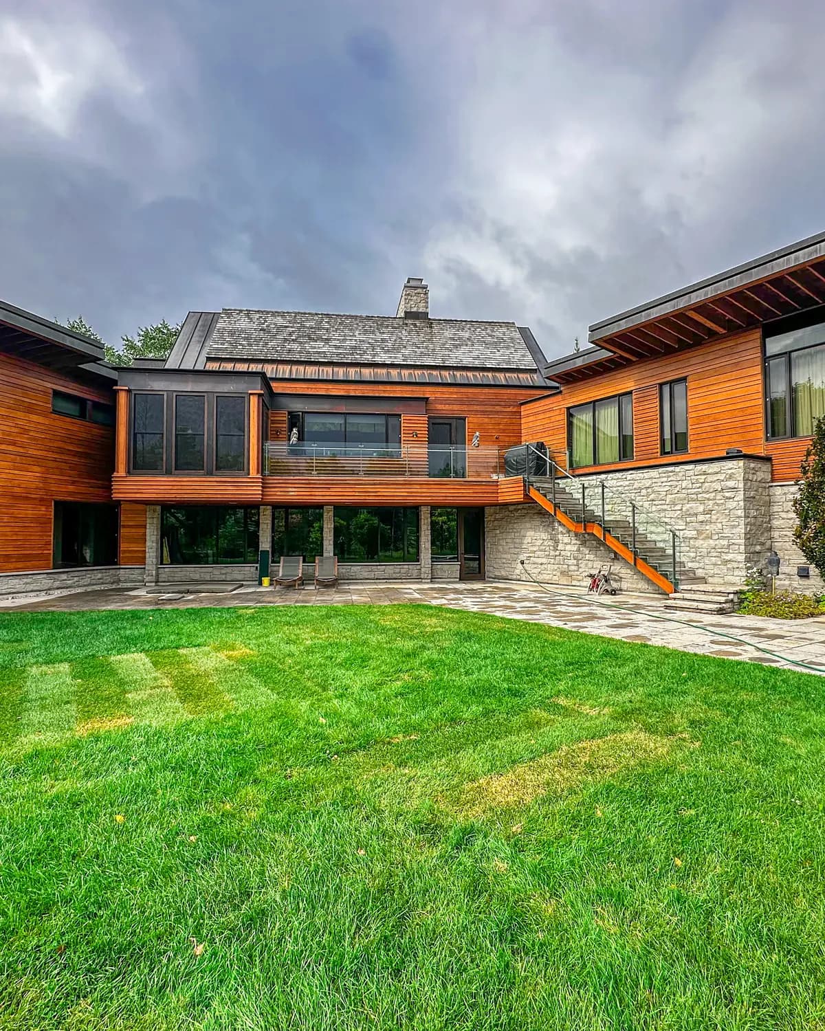 A modern Blue Mountain home with stained wood cladding and stone, seen across the lawn under a heavy sky.