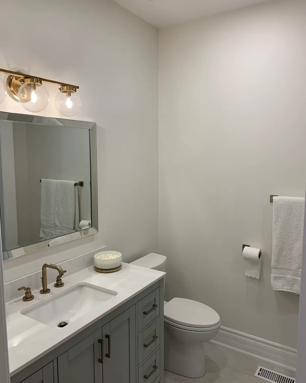 A powder room after painting, with a vanity, framed mirror and brass sconce against soft white walls.