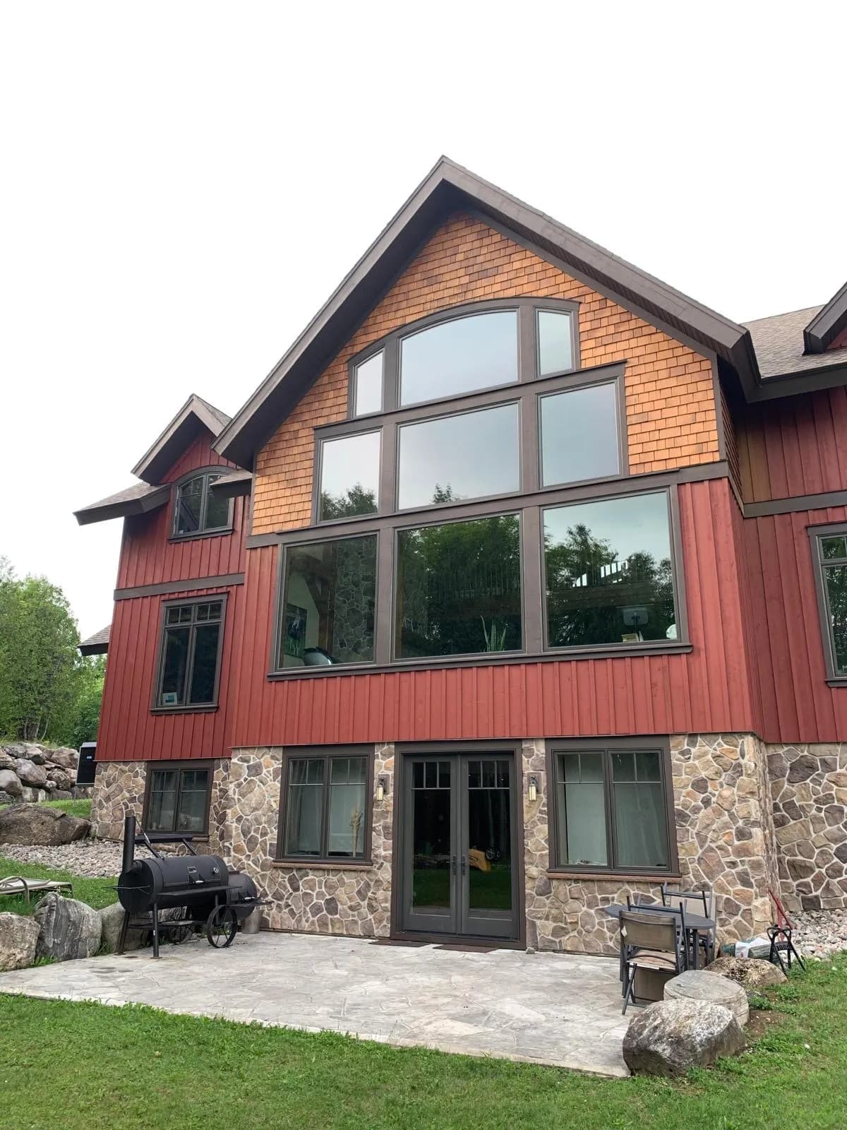 The window wall of a timber frame home near Markdale, restained cedar shakes in the gable above deep red board and batten and a stone base.