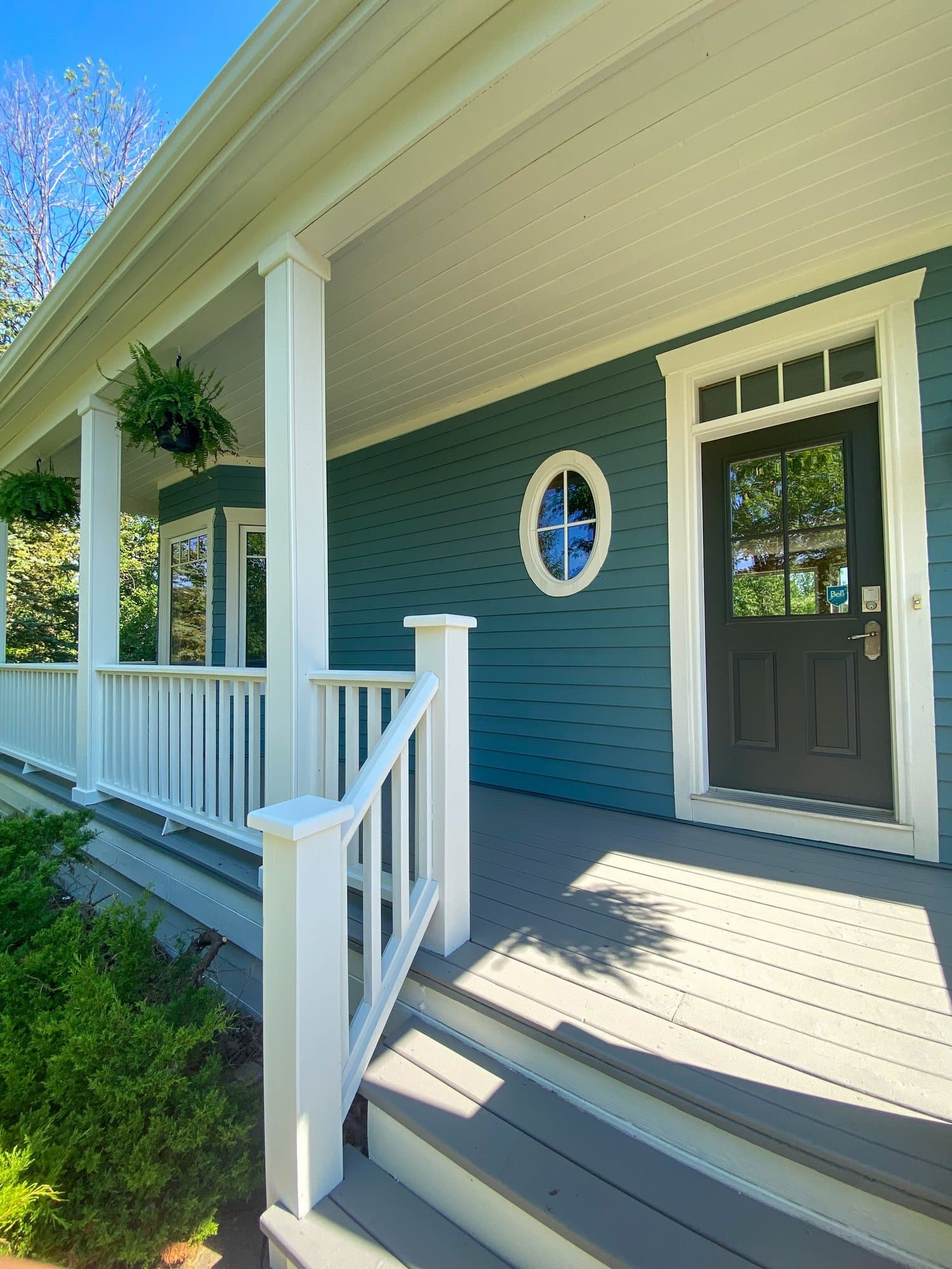 Freshly painted teal farmhouse porch with white columns, a grey deck and an olive green front door.