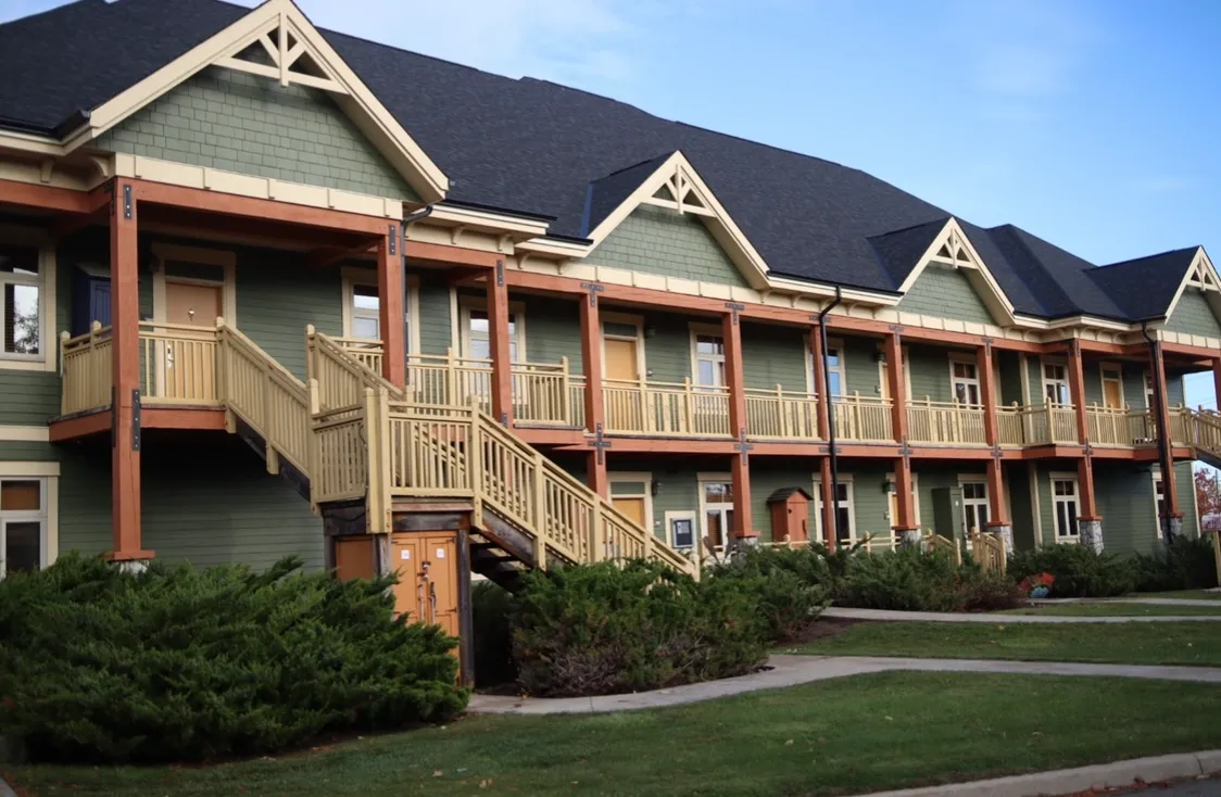 A lodge building at Hilton Grand Vacations Club Blue Mountain Canada with sage green siding, timber balconies and stairs finished in warm wood tones, and a dark shingle roof.